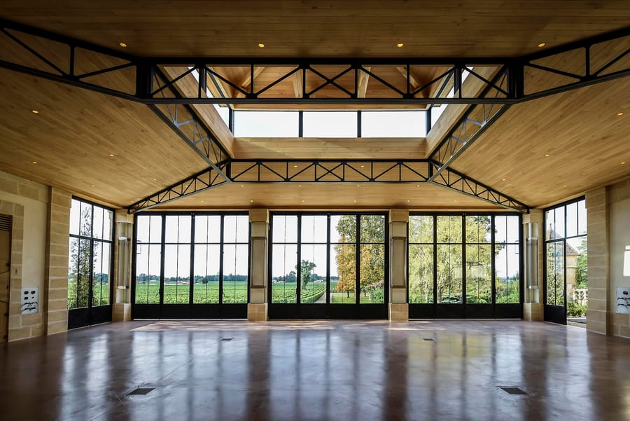 Grande salle contemporaine avec plafond en bois, charpente métallique et baies vitrées donnant sur des vignes, dans un domaine viticole, Château Beauregard à Pomerol