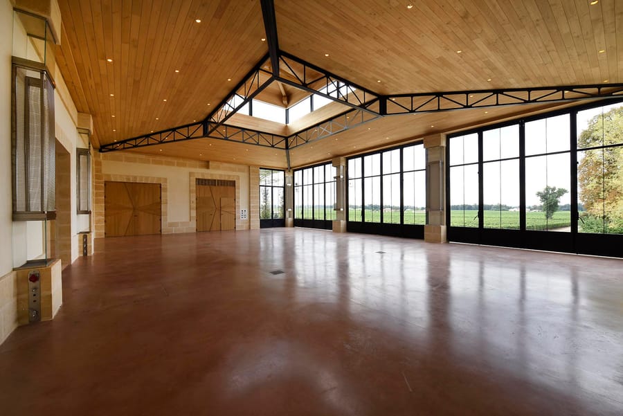 Vaste salle vide dotée d'un plafond en bois, de baies vitrées et d'une charpente métallique donnant sur les vignes, Château Beauregard à Pomerol