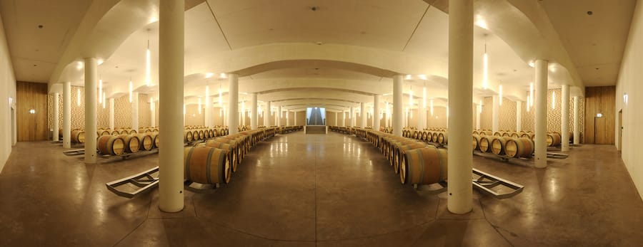 Alignement de barriques en bois sous des voûtes blanches éclairées dans un chai moderne, Château Cheval Blanc à Saint-Émilion