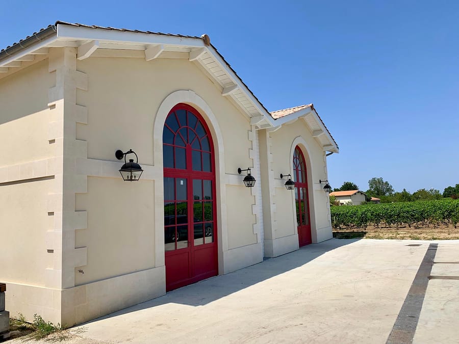 Bâtiment viticole aux murs clairs avec de grandes portes cintrées rouges et des lanternes noires, bordé par les vignes, Château de Côme à Saint-Estèphe
