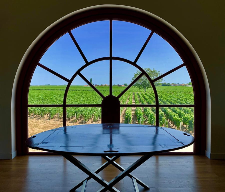 Table en bois sombre posée devant une grande fenêtre cintrée offrant une vue panoramique sur des vignes, Château de Côme à Saint-Estèphe