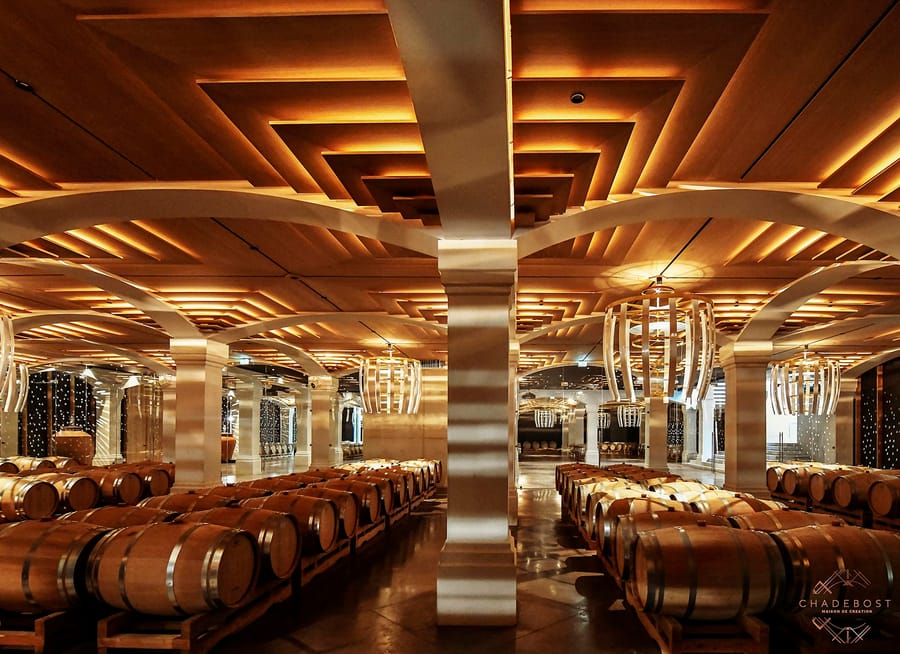 Barriques de vin alignées sous un plafond sculptural en bois dans le chai de Montlabert signé Chadebost