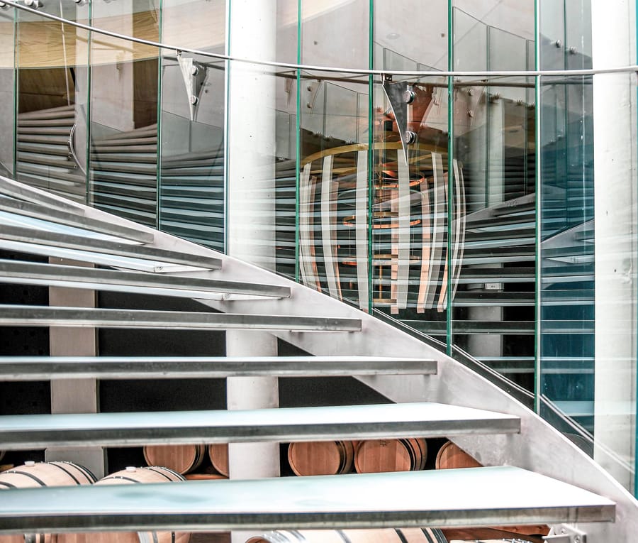 Escalier contemporain en métal et verre surplombant des fûts de chêne dans un chai moderne, Château Montlabert à Saint-Émilion