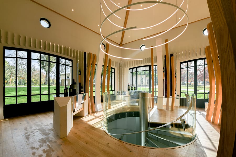 Intérieur d'une salle de dégustation moderne avec escalier en colimaçon, luminaire circulaire et baies vitrées donnant sur un parc, Château Montlabert à Saint-Émilion