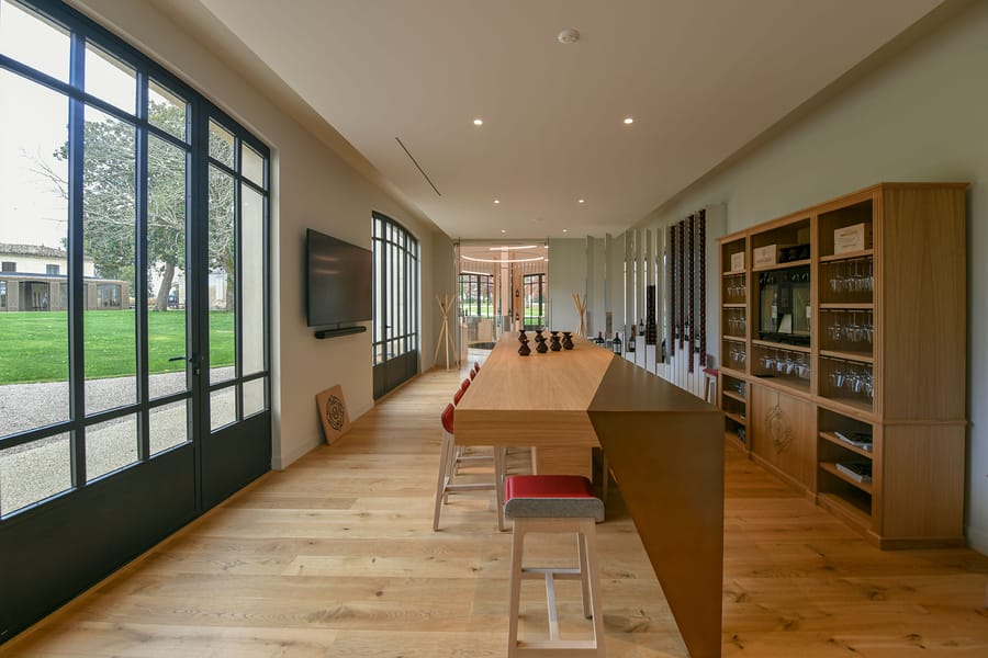 Salle de dégustation avec un long comptoir en bois, des tabourets et de grandes baies vitrées donnant sur un parc, Château Montlabert à Saint-Émilion