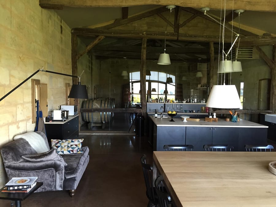Aménagement intérieur moderne avec canapé, cuisine ouverte et verrière donnant sur un chai à barriques, Château Tour du Pin à Saint-Émilion