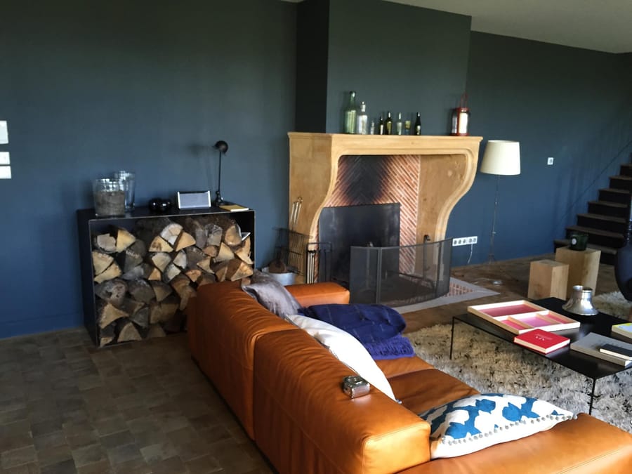 Salon chaleureux avec canapé en cuir marron, cheminée en pierre et meuble à bûches devant un mur bleu foncé, Château Tour du Pin à Saint-Émilion