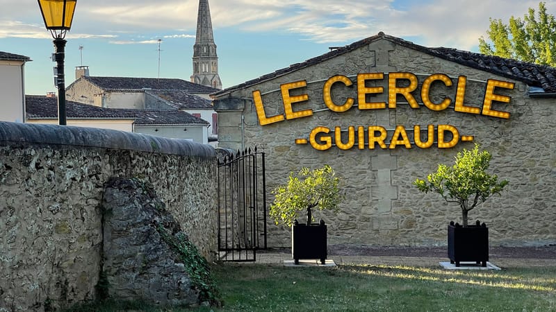 Façade en pierre du restaurant Le Cercle Guiraud avec son enseigne lumineuse jaune et un clocher en arrière-plan, à Sauternes