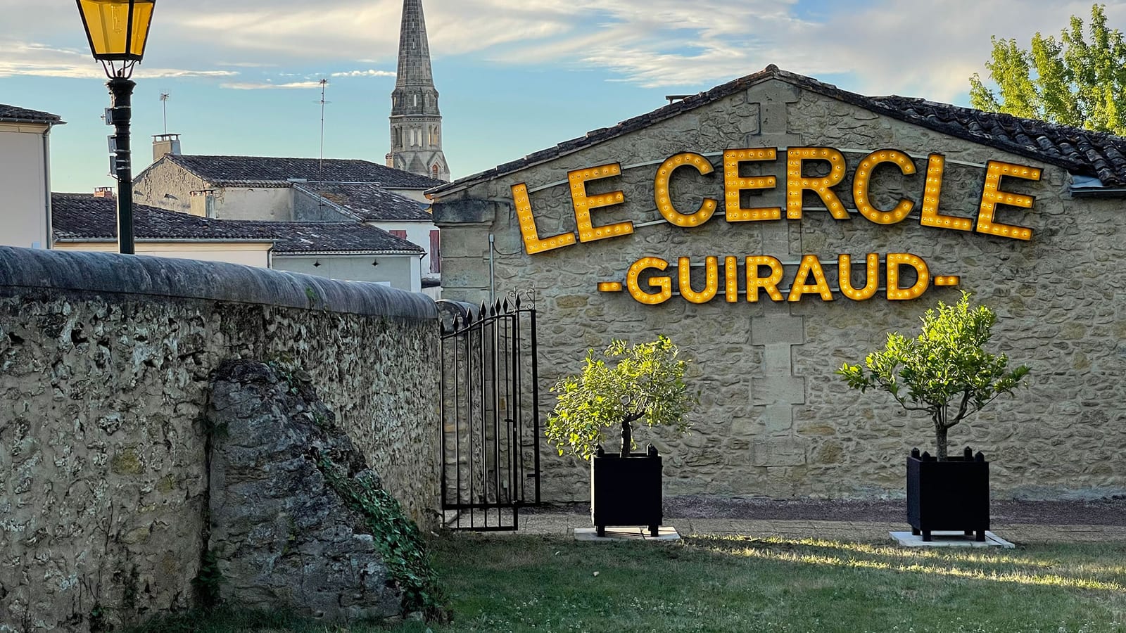 Façade en pierre du restaurant Le Cercle Guiraud avec son enseigne lumineuse jaune et un clocher en arrière-plan, à Sauternes