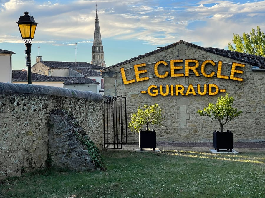 Façade en pierre du restaurant Le Cercle Guiraud illuminée par son enseigne jaune, avec deux arbustes en pot et un clocher en arrière-plan
