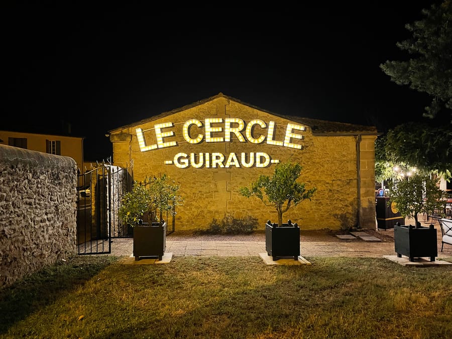 Façade en pierre illuminée de nuit du restaurant Le Cercle Guiraud avec son enseigne et des arbustes en pot, à Sauternes