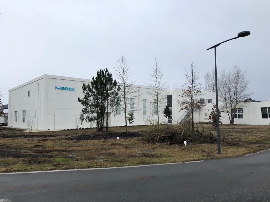 Façade d'un bâtiment moderne blanc portant le logo Merck et entouré de jeunes arbres, Merck Biodevelopment à Martillac