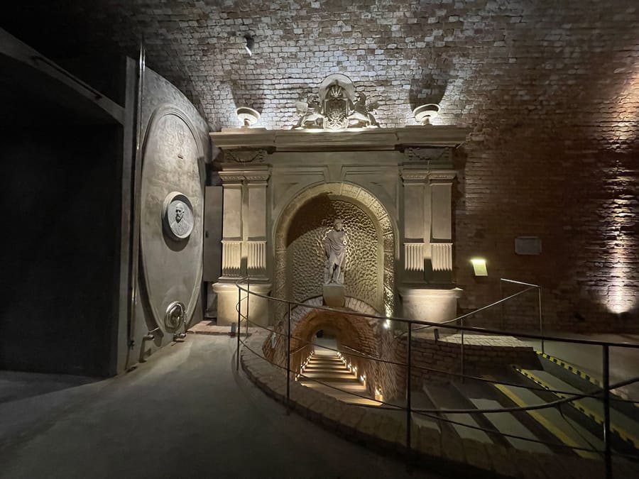 Cave voûtée en briques aménagée avec un escalier éclairé et un portail en pierre sculptée abritant une statue