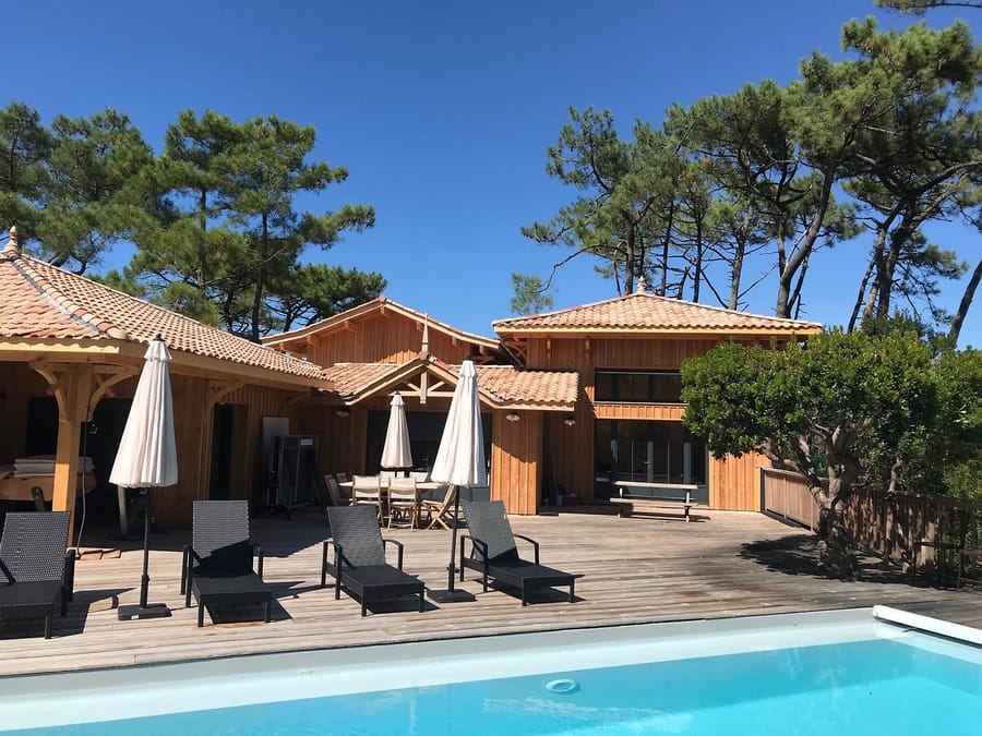 Terrasse en bois avec piscine et transats devant une villa en bois entourée de pins, au Cap Ferret